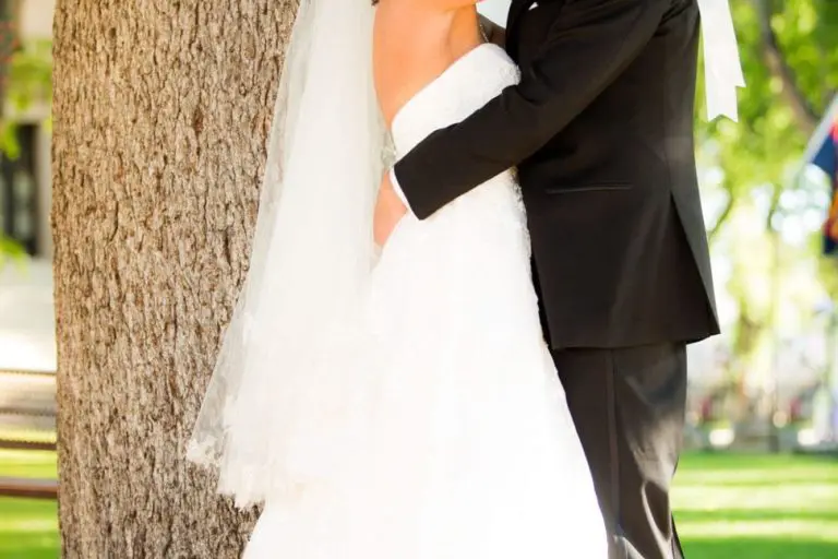 A bride in a white gown and veil smiles at the camera while hugging a groom in a black suit. They stand outdoors by a large tree, far from any seminar or corporate event, with sunlight filtering through green leaves.