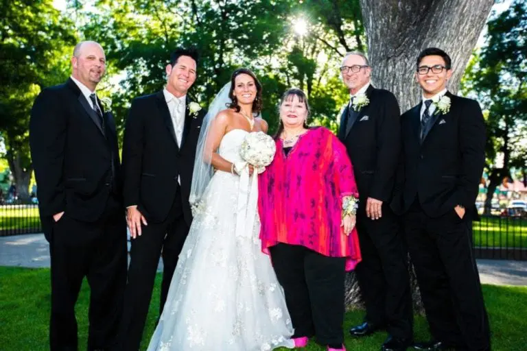 A bride in a white dress stands with five people, including men in black suits and a woman in a bright pink blouse, all smiling outdoors by a tree as if attending a joyful seminar in a sunlit park.