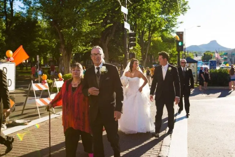 A bride and groom cross a sunlit street with two older adults, one using a cane. Dressed formally, they pass trees, barricades, and festive decor—evoking the elegance of a corporate event outside the seminar hall.