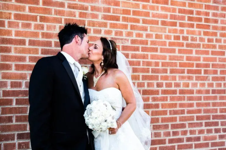 A bride and groom in wedding attire share a kiss in front of a red brick wall, bringing romance to the site of a recent corporate event. The bride holds white flowers and wears a strapless dress, while the groom is dressed in a black suit.