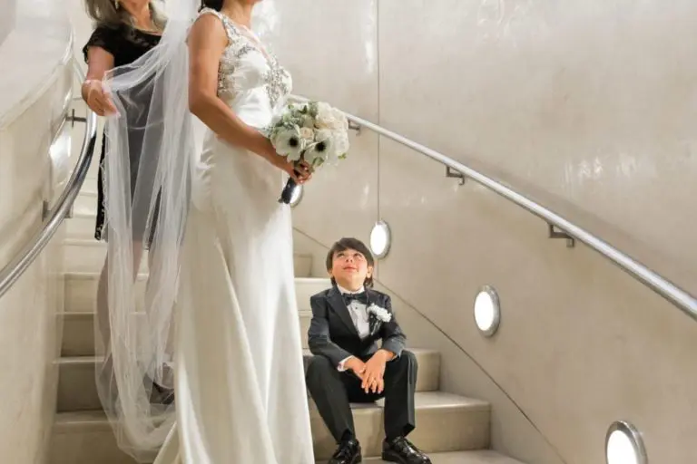 A bride in a white gown holding a bouquet stands on a curved staircase, smiling, with an older woman behind her and a young boy in a tuxedo on the step below. The modern setting with tall vertical lights resembles a chic corporate event venue.