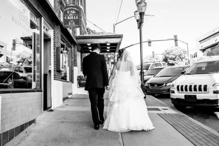 A bride in a wedding dress and veil walks down a city sidewalk holding hands with a man in a suit, as if heading to a corporate event, passing cars parked along the street on a bright day.