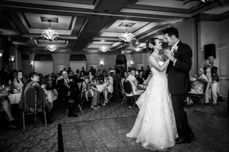 A bride and groom share their first dance in a ballroom, elegantly lit and decorated as if set for a corporate event. Seated guests watch the smiling couple—the bride in white, the groom in a dark suit—enjoy this unforgettable moment.
