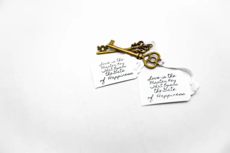 Two vintage-style brass keys with heart-shaped bows are attached to white tags. Perfect as unique favors for a corporate event or seminar, the tags read, "Love is the master key that opens the gate of happiness," on a plain white background.
