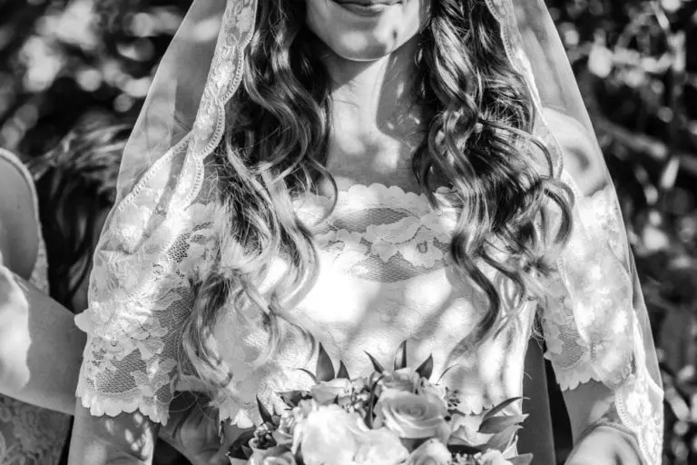 A bride in a lace dress and veil stands outdoors, holding a bouquet of roses. Sunlight and shadows dance across her face and hair as she smiles gently, the greenery behind her creating a serene backdrop—far from any seminar or corporate event.
