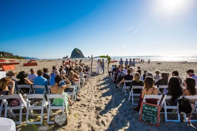 A wedding ceremony is taking place on a sandy beach under a clear blue sky, with guests seated in white chairs facing an arch. Haystack Rock is visible in the background near the ocean shoreline, creating an elegant setting perfect for any seminar or special occasion.