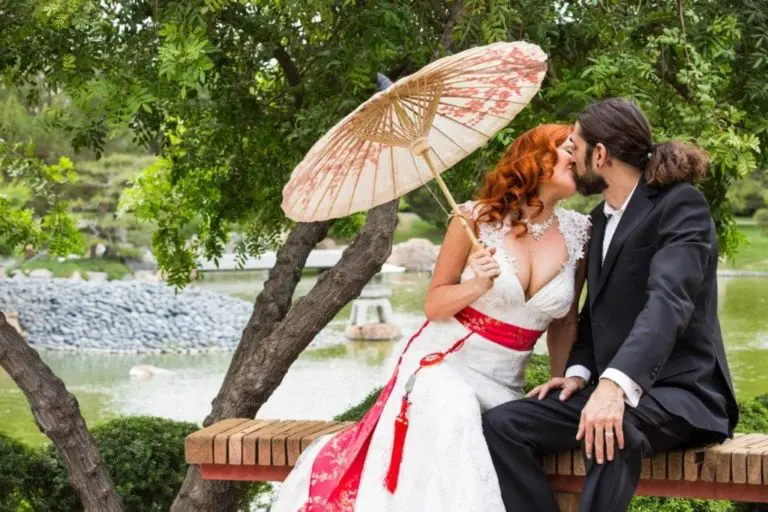 A bride in a white dress with a red sash and a groom in a black suit share a kiss under a parasol. They sit on a bench by a serene pond, surrounded by lush greenery. This magical moment showcases the essence of event photography.