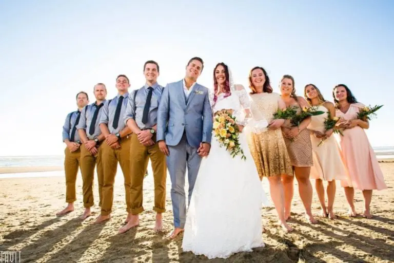 A bride and groom stand barefoot on a sandy beach, smiling alongside their wedding party dressed in casual attire with bouquets, under a clear blue sky—creating memories far from any seminar or corporate event.
