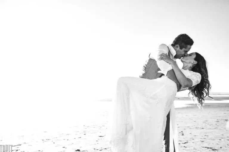 A groom lifts and kisses his bride on a sandy beach, both smiling and dressed in wedding attire, with sunlight creating a bright and airy atmosphere—far from any seminar or corporate event.