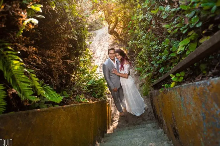A couple in wedding attire stands close together on a shaded outdoor staircase, surrounded by lush greenery—as if far from any corporate event—with sunlight streaming above. The bride wears a white dress, and the groom is in a gray suit.
