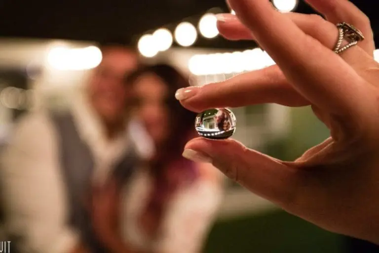 A hand with a ring holds a small glass sphere, reflecting an image of a blurred couple in the background at a seminar, with glowing string lights above them.