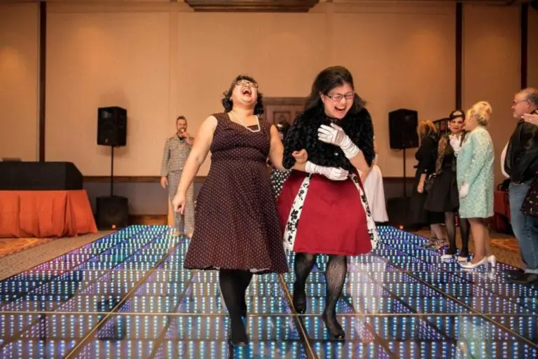 Two women in vintage-style dresses laugh and walk arm in arm across a colorful, illuminated dance floor at a corporate event, while other people stand and watch in the background.