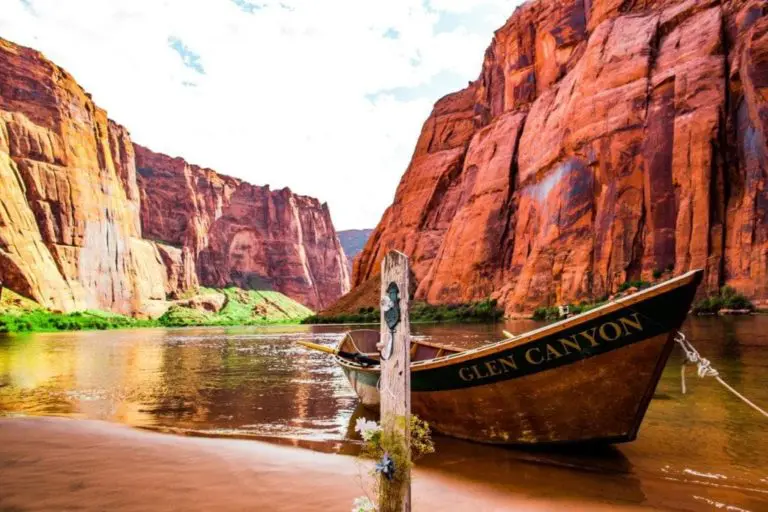 A wooden boat labeled "Glen Canyon" is tied to a post on the calm Colorado River, its peaceful setting framed by towering red rock cliffs—an inspiring backdrop for a seminar or corporate event under a bright, partly cloudy sky.