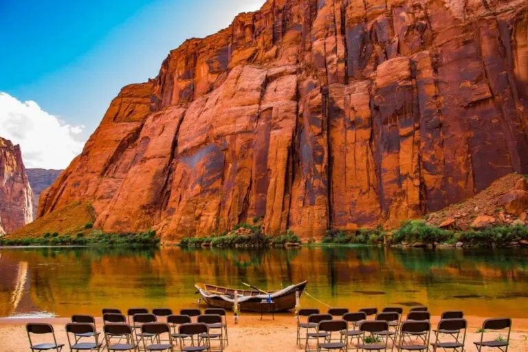 Rows of empty chairs are arranged for a seminar or corporate event, facing a small boat docked on a calm river, framed by towering red rock cliffs under a partly cloudy sky.