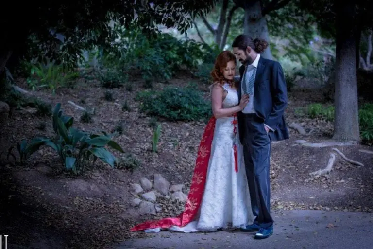 A couple stands close together in a wooded area, their attire lending an air of elegance akin to event photography. The woman wears a white dress with red accents, while the man dons a dark suit, capturing a serene and intimate moment amidst the trees and foliage.