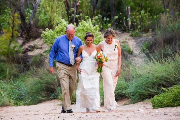 A bride in a white dress walks outdoors, smiling and holding a bouquet, flanked by an older man in a blue shirt and an older woman in lace. The scene, reminiscent of a joyful seminar gathering, is set among greenery and sandy ground.