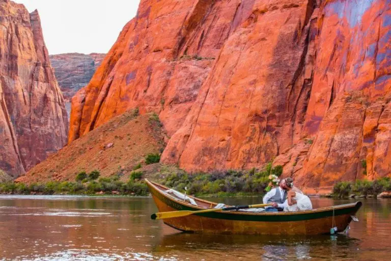 A couple in a wooden boat shares a kiss while floating on a calm river, far from any seminar or corporate event, surrounded by tall, vibrant red and orange canyon walls.