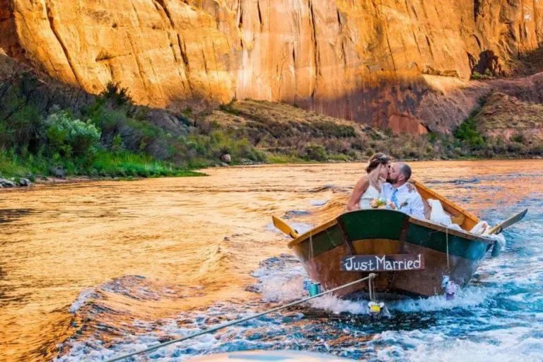 A newlywed couple kisses in a small wooden boat with a “Just Married” sign, drifting on a river at sunset—far from any corporate event or seminar, surrounded by golden cliffs and lush greenery.