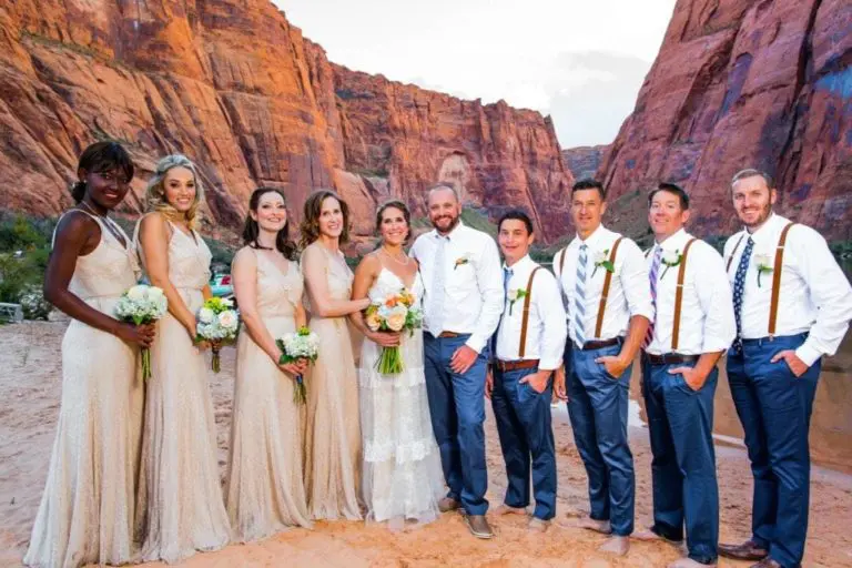 A wedding party of five women in beige dresses and five men in white shirts with suspenders and ties pose together on sandy ground, as if ready for a seminar, with tall red rock cliffs rising in the background.