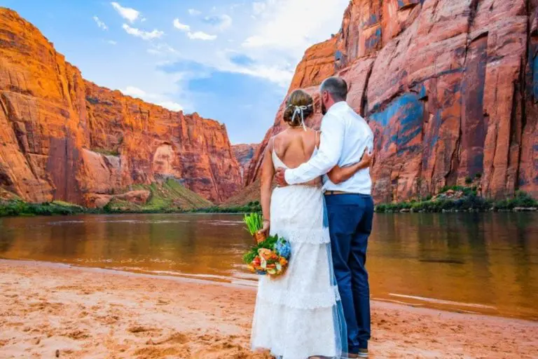 A bride and groom stand arm in arm on a sandy riverbank, facing dramatic red rock cliffs and a serene river, as if taking a moment away from a busy seminar or corporate event; the bride holds a colorful bouquet.