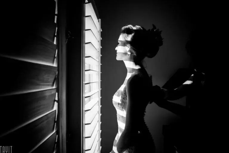 A woman in a bridal gown stands in profile by a window with horizontal blinds, sunlight casting dramatic stripes across her face and body, as another person adjusts her dress before a seminar or corporate event in the background.