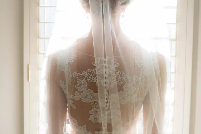 A bride in a lace wedding dress and floral headpiece stands with her back to the camera, gazing out a sunlit window through white blinds, as if quietly reflecting before a seminar or corporate event begins.