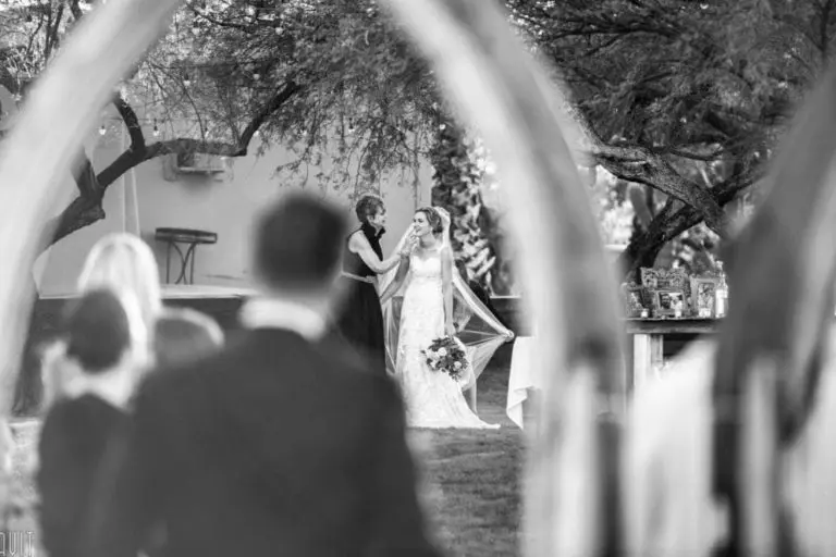 A black-and-white photo captures a bride holding a bouquet and smiling as a woman adjusts her hair. The scene, framed by blurred figures in the foreground, evokes the candid atmosphere of a seminar or corporate event audience.
