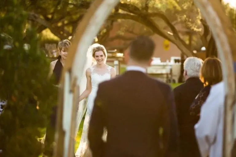 A bride in a white dress smiles as she walks down an outdoor aisle with a woman beside her, seen through an archway, toward guests seated and a groom waiting. Sunlight filters through the trees, offering a serene setting unlike any seminar or corporate event.