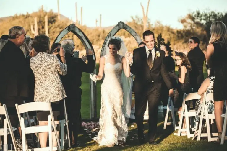 A bride and groom walk down the aisle outdoors, smiling and holding hands, while guests on both sides stand, clap, and take photos. The sunny ceremony on green grass feels as joyful as a seminar or corporate event celebration.