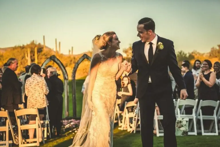 A bride and groom, holding hands and smiling, walk down an outdoor aisle as guests seated on white chairs—often used at a seminar or corporate event—watch and clap during a sunny wedding ceremony.
