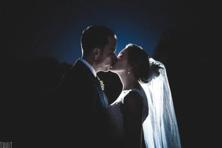 A bride and groom share a romantic kiss in low light, with soft backlighting highlighting the bride’s veil and their silhouettes—capturing an intimate moment far removed from the bustle of a corporate event or seminar.