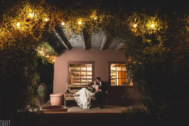 A bride and groom sit closely together on a porch bench at night, surrounded by greenery and warm string lights, with a softly lit window behind them—capturing an intimacy far from any corporate event or seminar.