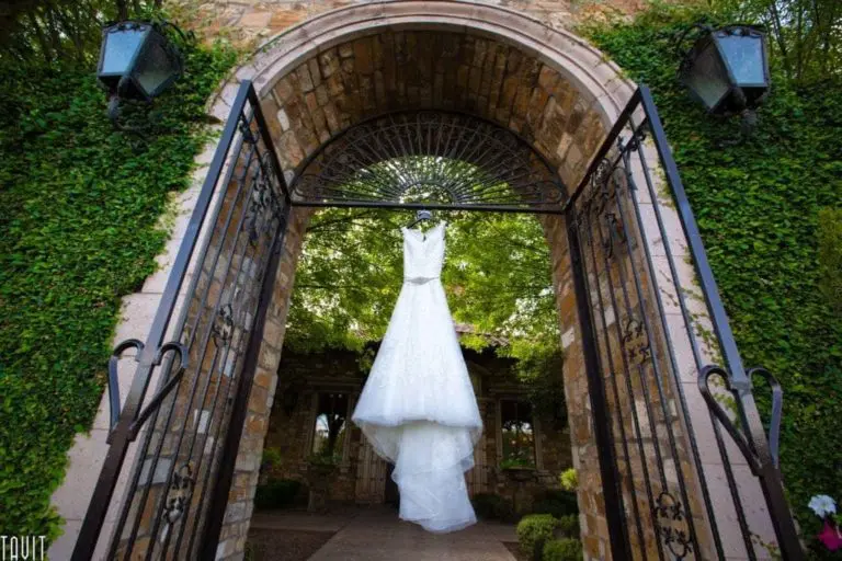 A white wedding dress is elegantly hanging on a wrought iron gate, framed by stone walls and green ivy, embodying the enchantment of event photography. In the background, a garden with trees and a stone building creates an idyllic setting, while two large lanterns grace the gate pillars.