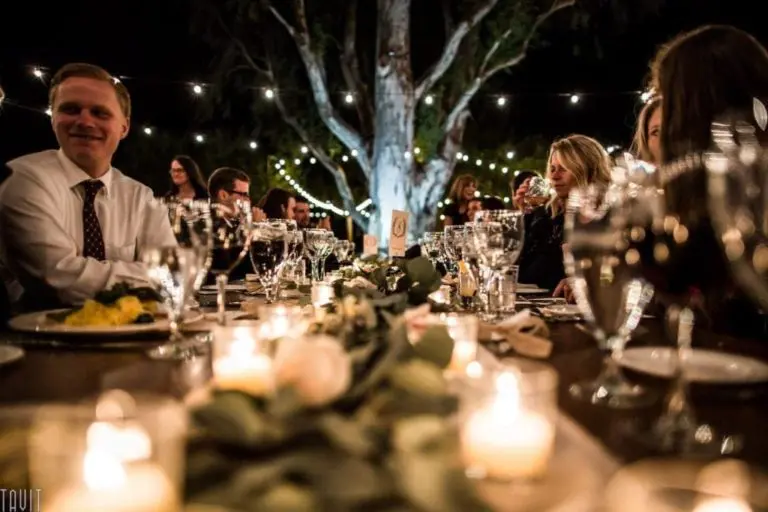 People sit around a long, beautifully decorated table outdoors at night during a corporate event, with string lights above, candles on the table, and trees in the background, creating a warm, festive atmosphere.
