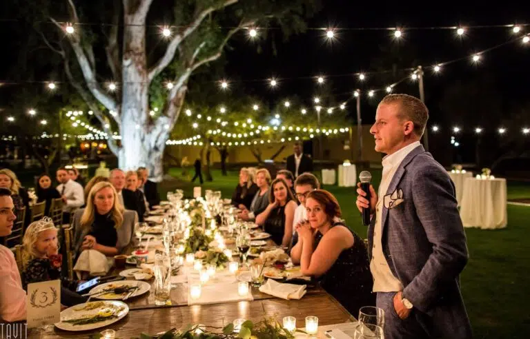 A man in a suit gives a speech with a microphone at an outdoor corporate event, as guests seated at a long, decorated table with candles listen attentively under string lights.