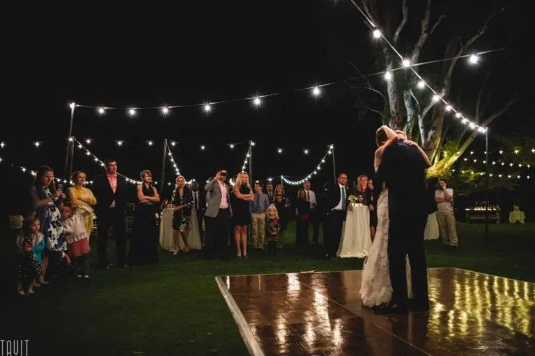A bride and groom share their first dance at night under string lights, while guests—much like at a corporate event or seminar—gather on the grassy outdoor dance floor, capturing the magical moment in photos.