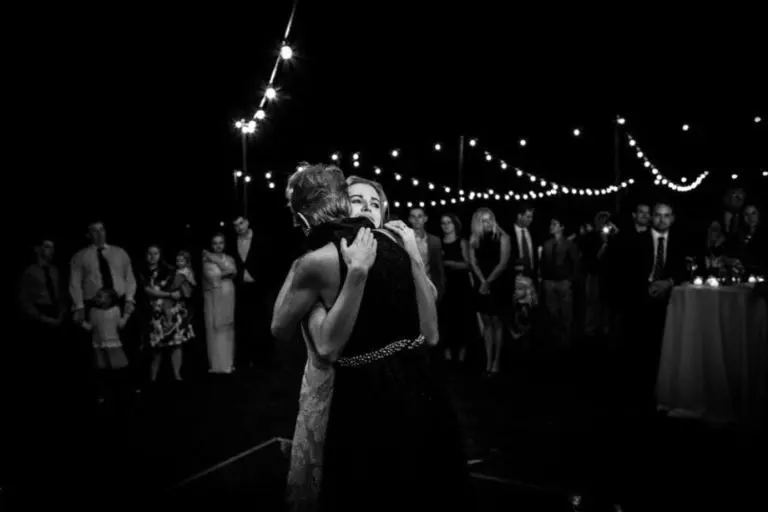 A bride and an older woman embrace on a dance floor at night, surrounded by guests under string lights, during an outdoor wedding celebration—not your typical corporate event or seminar setting.