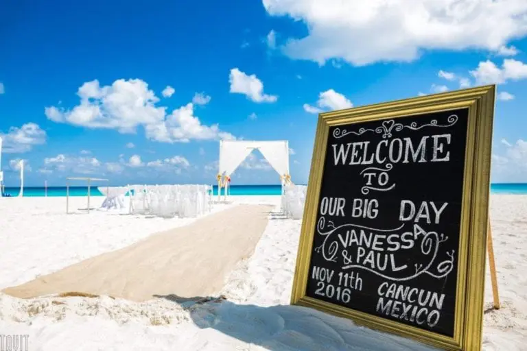 A chalkboard sign on a sandy beach reads "Welcome to our big day, Vanessa & Paul, Nov 11th 2016, Cancun, Mexico." In the background, a wedding arch and chairs are set up near the turquoise ocean—far from any seminar or corporate event.