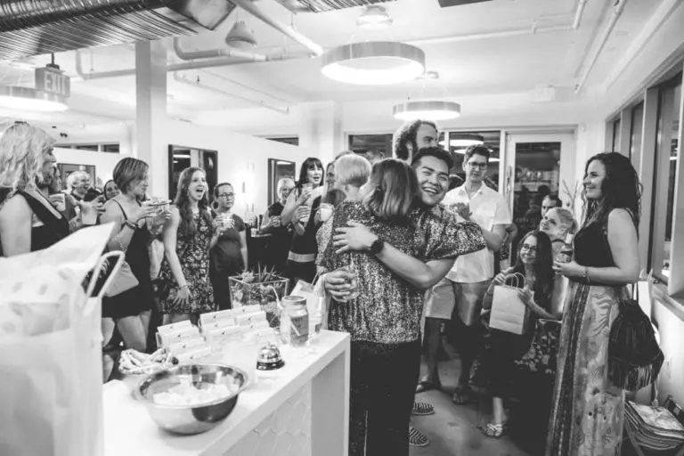 A black and white photo of a lively indoor corporate event. Two people hug in the center while others stand around smiling, laughing, and watching. The setting appears festive, with gifts and treats on a counter in the foreground.