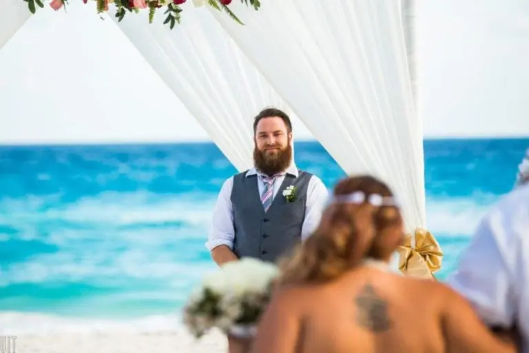 A bearded man in a vest and tie stands under a white draped arch on a beach, smiling as he waits for a seminar or corporate event to begin. The ocean is in the background, with blurred guests in the foreground.