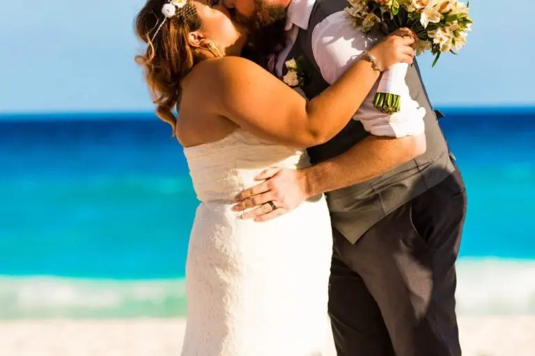 A newlywed couple kisses under a white canopy on a beach, with the ocean and blue sky in the background. Their romantic moment feels worlds away from any corporate event or seminar, as the bride wears a lace dress and flower crown, holding her bouquet.
