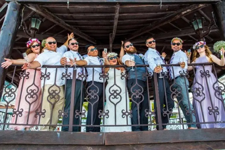 A wedding party of eight people stands on a balcony, smiling and holding drinks. Their festive attire and sunglasses bring a lively touch—more celebration than seminar or corporate event, with purple dresses, one woman in white, and men in shirts and pants.
