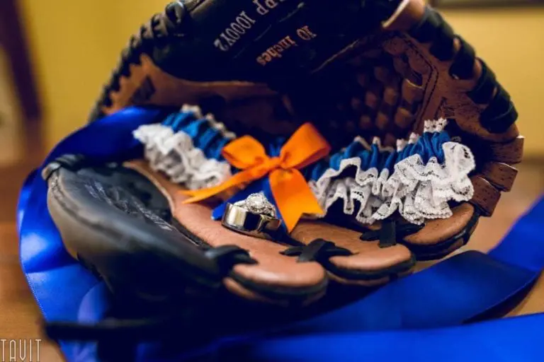 A close-up of a baseball glove holding a wedding garter with blue, white, and orange ribbon, alongside two wedding rings and blue ribbon accents—perfect as inspiration for a unique seminar or corporate event celebration.