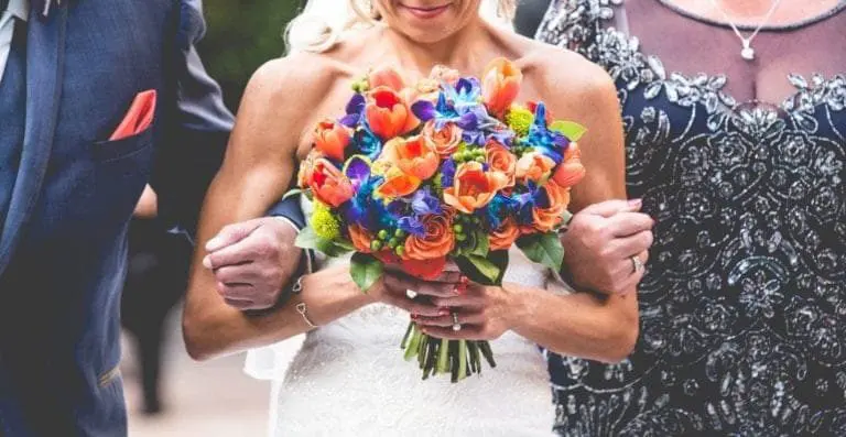 A bride in a white dress holds a colorful bouquet of orange and blue flowers while walking arm-in-arm with two well-dressed people, as if arriving at an elegant seminar or corporate event.