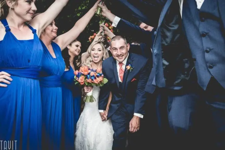 A bride and groom smile joyfully while walking hand-in-hand through a tunnel formed by wedding party members in blue dresses and suits, celebrating with the excitement of a successful seminar. The bride holds a vibrant bouquet.