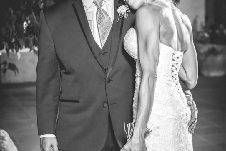 A bride in a lace gown rests her head affectionately on the groom’s chest as he looks down. She holds a bouquet, and both appear content—as if sharing a quiet moment after a seminar or corporate event. Outdoors at night, softly lit. Black-and-white photo.
