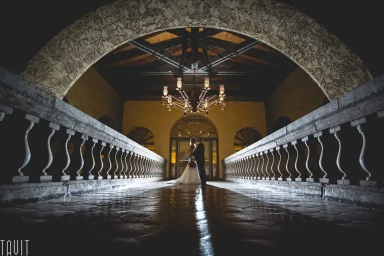 A bride and groom stand together at the center of a stone bridge under a chandelier, framed by an arched ceiling and dramatic lighting—an enchanting setting that could inspire any seminar or corporate event with its romantic atmosphere.