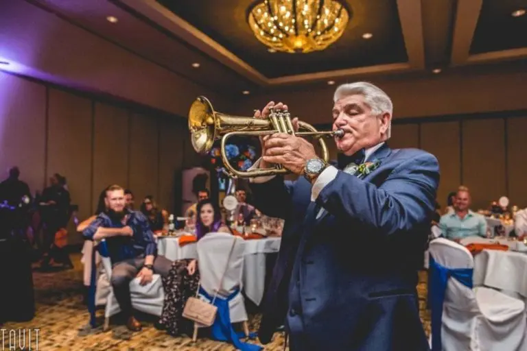 An older man in a navy suit plays the trumpet at an indoor corporate event, with guests seated at decorated tables and a chandelier hanging above. People watch and smile, creating a festive atmosphere.