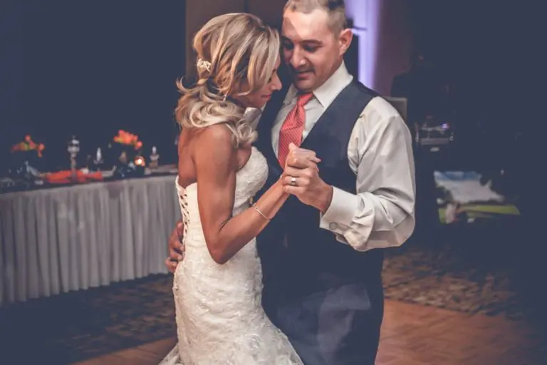 A bride in a white lace gown and a groom in a vest and tie share a slow dance on a wooden floor at their wedding reception, transforming the venue from seminar or corporate event space into a romantic setting, with a decorated table visible in the background.