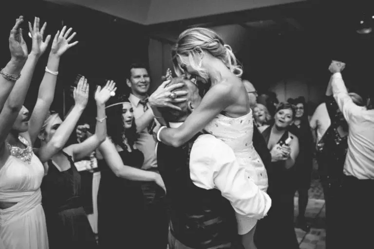 A black and white photo of a joyful celebration, showing a man lifting a woman in a crowd of people with their hands raised, dancing and smiling at what appears to be a festive corporate event or wedding reception.